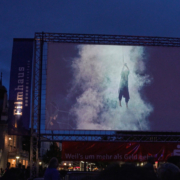 Eine bespielte Leinwand vor dunkelblauem Himmel, im Hintergrund das Theater Bielefeld.
